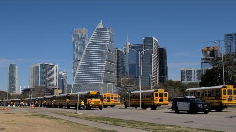 Photo of several tall glass-clad buildings. The one in front has an ascending curve that makes it sail-shaped. For some reason, there are two lines of school buses and a police vehicle in the parking lot in the foreground.