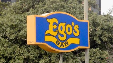 Photo of a sign on a pole in front of treetops that reads "Ego's Bar."
