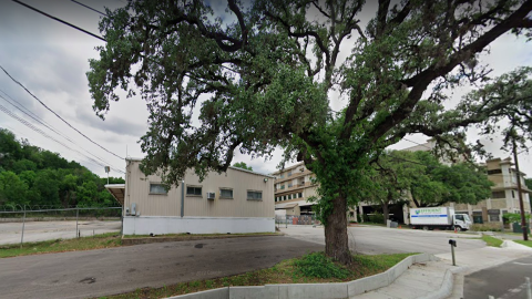 Photo of a concrete parking lot with a one-story metal building on it and a large tree in front.