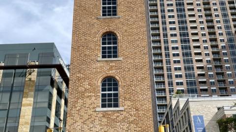 A narrow, six-story brick tower built in the 1930s with modern midrise and high-rise buildings behind it.