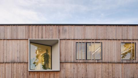 Photo of a section of a two-story flat-roofed building clad in wood with an angled and regular window.